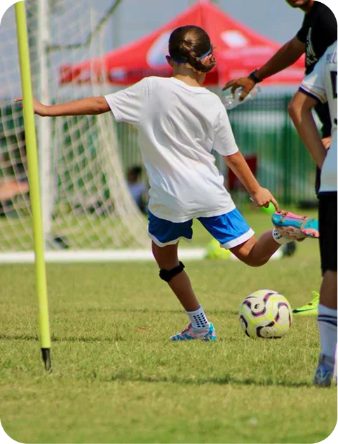 Child wearing a white shirt and blue shorts kicking a soccer ball on a grassy field near a goalpost.