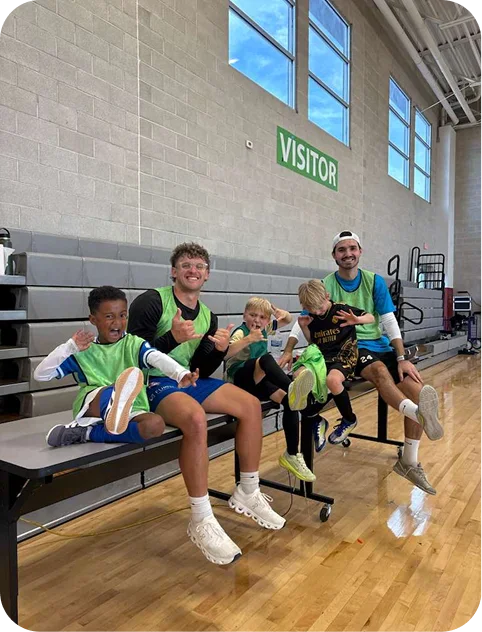 Two adult men and three boys sitting on bleachers in a gym, all wearing green sports pinnies, posing playfully with thumbs up and animated expressions.
