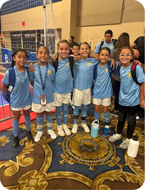 Six young girls in light blue soccer uniforms standing arm in arm indoors with adults and a net in the background.