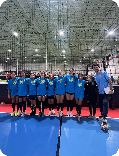Indoor group photo of a youth girls soccer team in blue shirts with a coach to the right and a soccer ball on the floor.