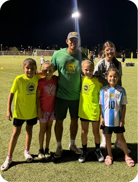 Youth soccer coach posing at night on a field with five smiling children wearing soccer jerseys and casual sportswear.