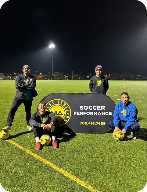 Four men posing on a soccer field at night around a sign that says 'Next Level Athletics Soccer Performance' with a phone number, each holding a yellow soccer ball.
