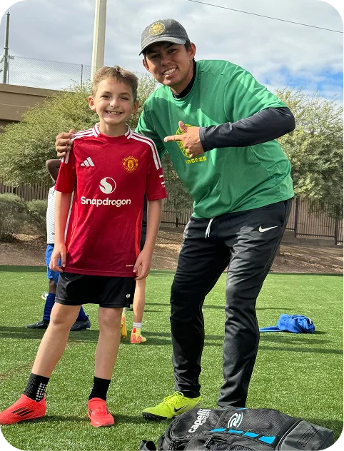 Smiling boy in red Manchester United soccer jersey standing next to a man in a green shirt and black pants pointing at him on a grassy field.