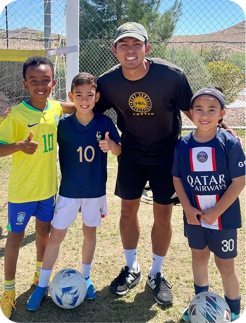 Three boys in soccer jerseys and a man in black sportswear smiling outdoors on a sunny day near a soccer goal, each boy standing with a soccer ball.
