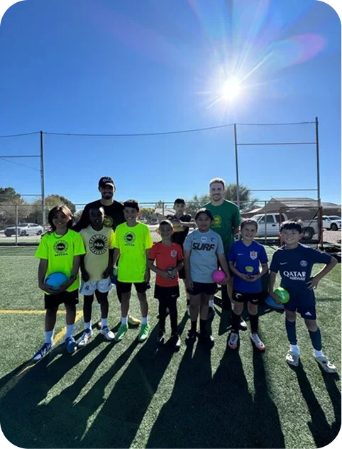 Group of young boys and two coaches on a sunny soccer field, some holding colorful balls.