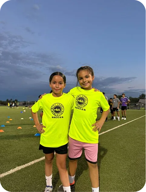Two young girls in bright yellow soccer shirts standing on a soccer field with arms around each other during evening practice.