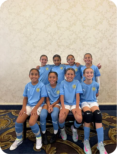 Group of eight young girls in matching light blue soccer uniforms posing indoors on a patterned carpet against a beige floral wallpaper.