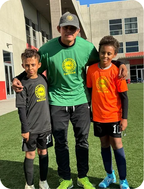 Coach in green shirt and cap standing with two boys in black and orange soccer uniforms on a grassy field.