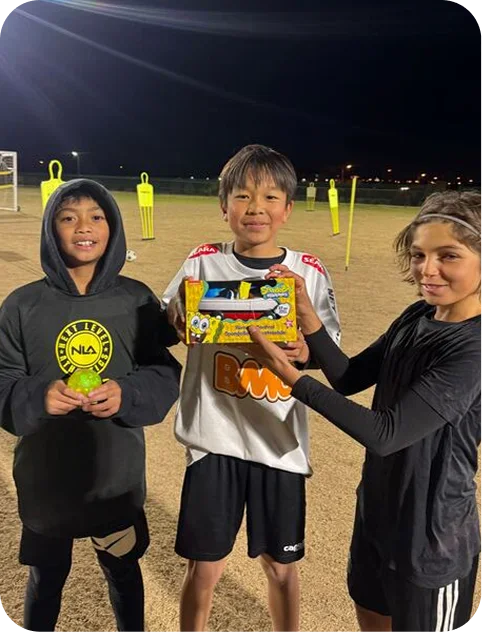 Three children standing on a soccer field at night, holding a SpongeBob SquarePants toy box and a small yellow trophy, with training mannequins in the background.