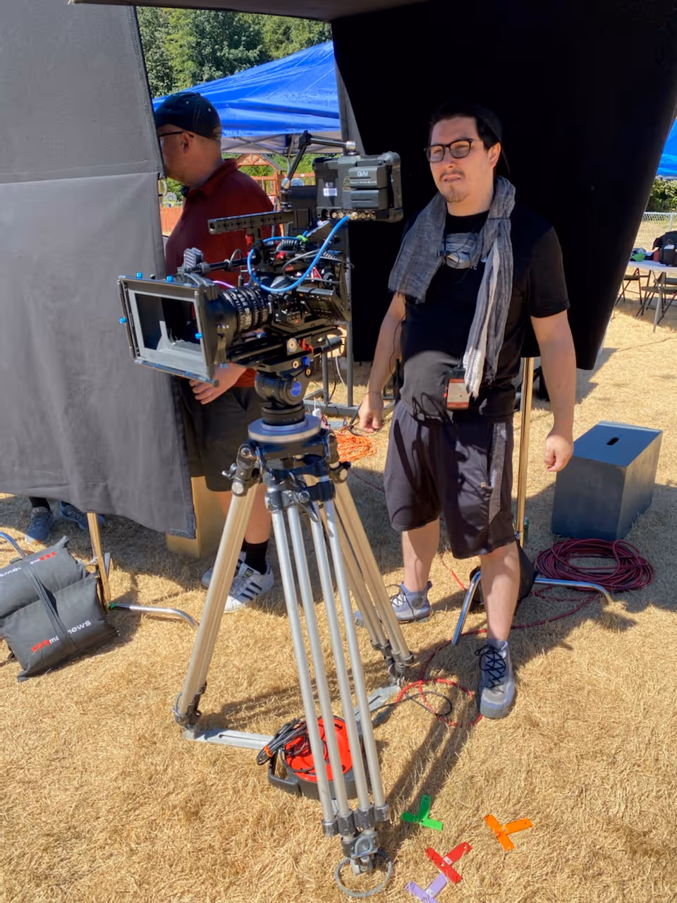 Man wearing glasses, a scarf, and black clothes stands next to a professional film camera mounted on a tripod outdoors on dry grass.