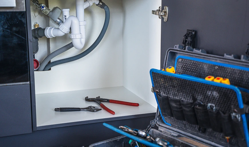 Open keukenkastje onder de gootsteen met zichtbare afvoerbuizen, handgereedschap in de kast en een open gereedschapskoffer op de vloer.
