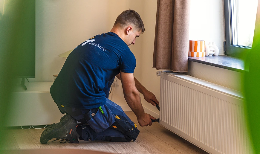 Monteur knielt bij een radiator en voert werkzaamheden uit aan de verwarming in een woonruimte.