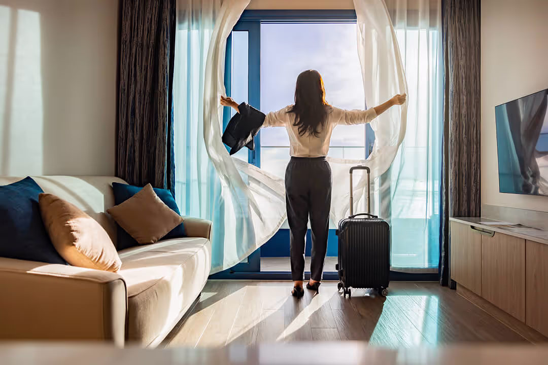 Frau steht mit Rollgepäck in einem Hotelzimmer und öffnet die Vorhänge zu einem Balkon mit Tageslicht.