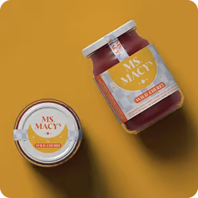 Two jars of Ms. Macy's Wild Cherry jam on a mustard yellow background, one standing upright and one lying on its side.