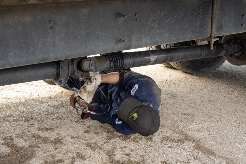 Technician lying under a truck working on the drive train with a grease gun and protective gloves.