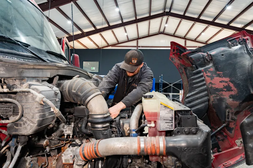 Technician performing preventive maintenance on a semi-truck engine inside a service bay with hood raised.
