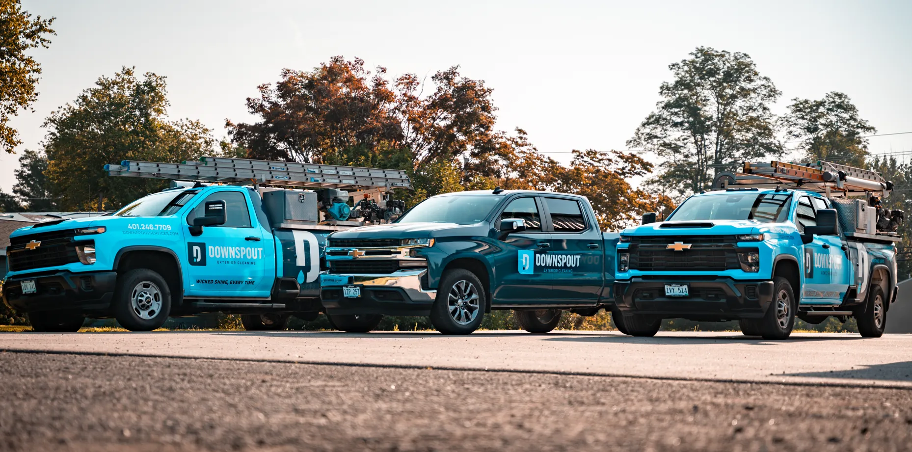 Three Downspout Exterior Cleaning trucks parked side by side outdoors with ladders on top, under clear sky and trees.