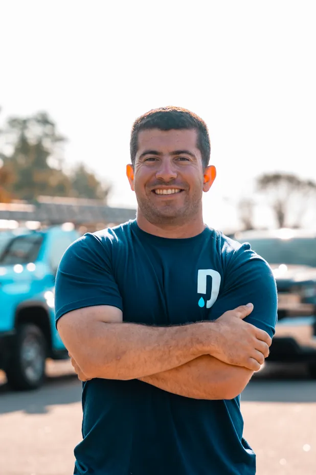 Smiling man with short dark hair and muscular arms standing with crossed arms outdoors in front of parked vehicles.