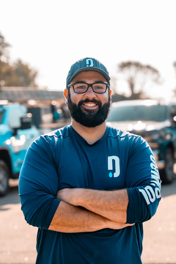 Bearded man with glasses smiling, wearing a blue cap and long-sleeve shirt with a logo, standing outdoors with crossed arms.