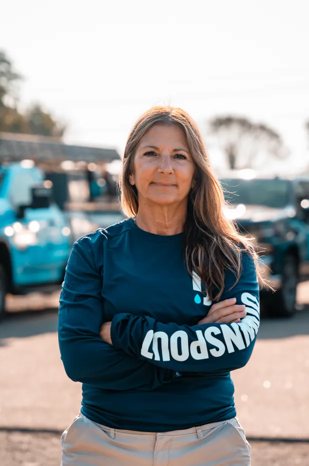 Confident woman with long hair wearing a blue long-sleeve shirt with 'JOHN'S POUT' text, standing outdoors with arms crossed.
