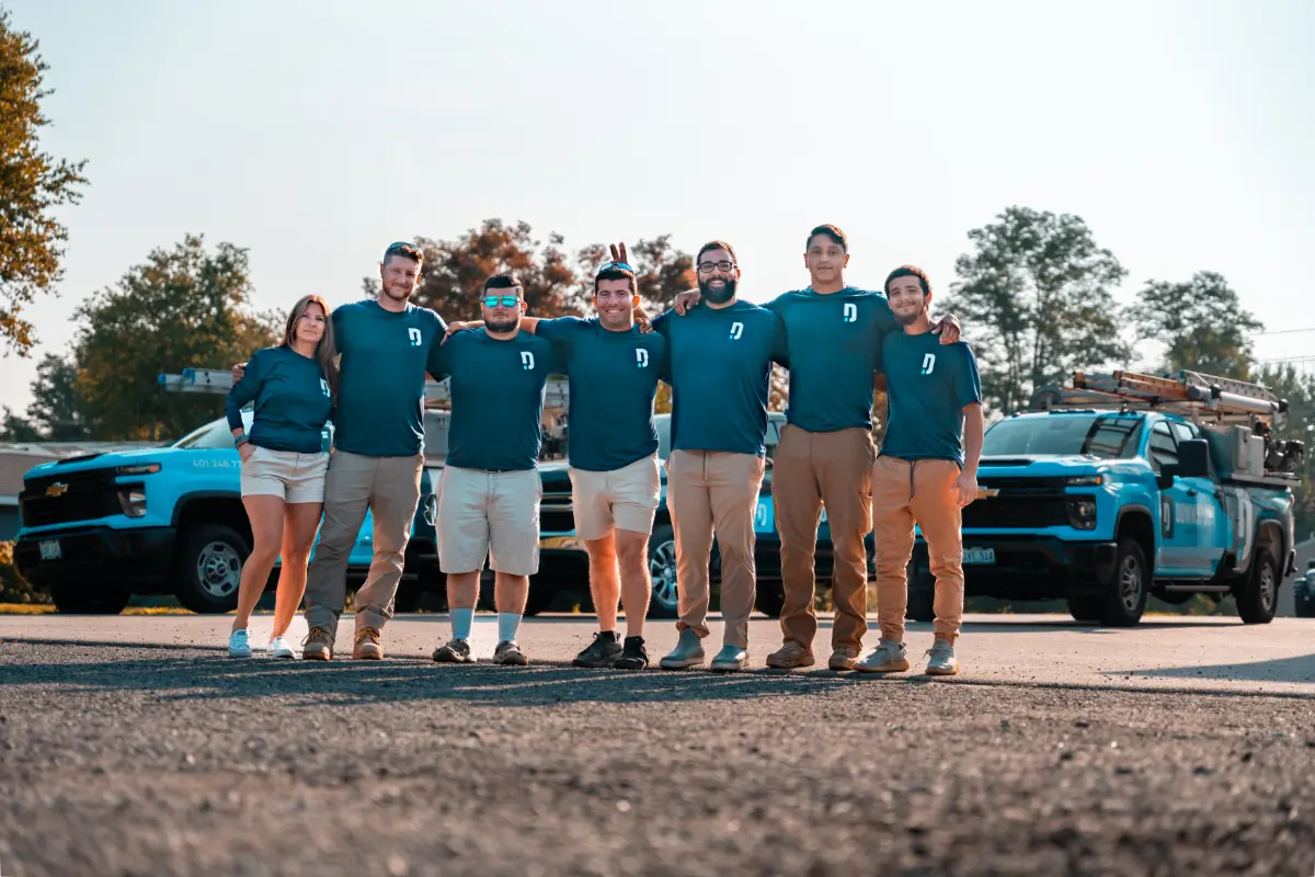 Group of seven people in matching teal shirts standing side by side with arms around each other in front of blue company trucks on a sunny day.