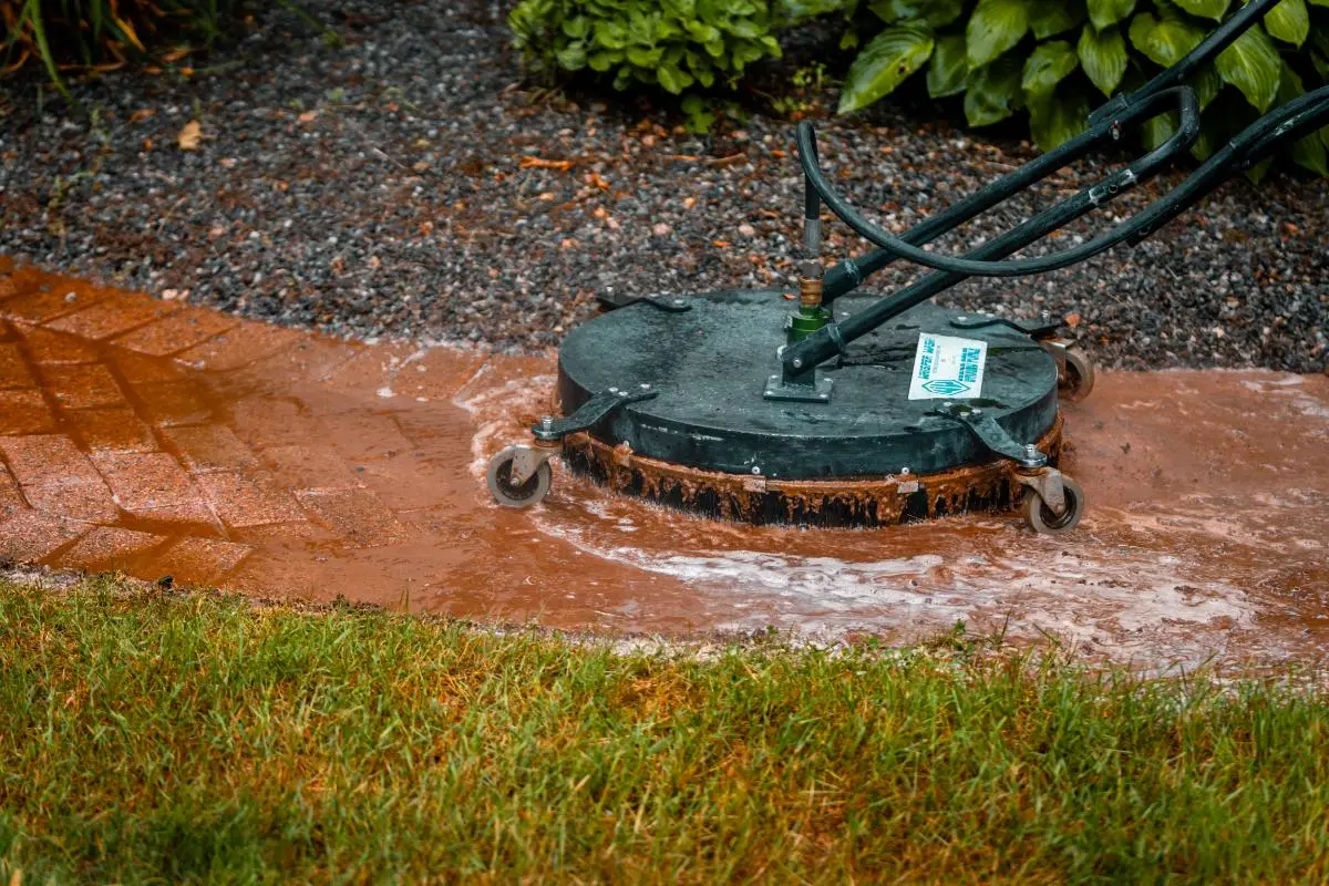 Surface cleaner machine spraying water on brick pavers near grass and gravel.