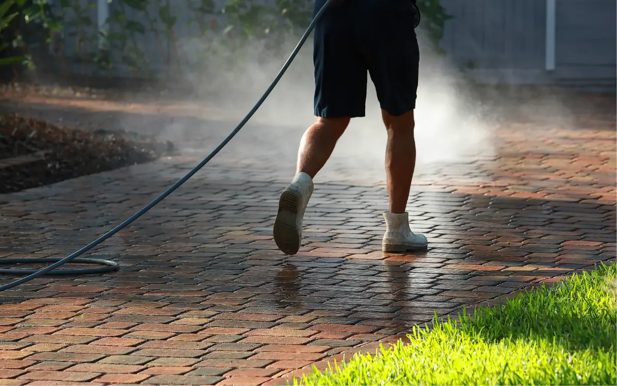 Person wearing boots and shorts power washing a brick paved driveway with water spray visible.