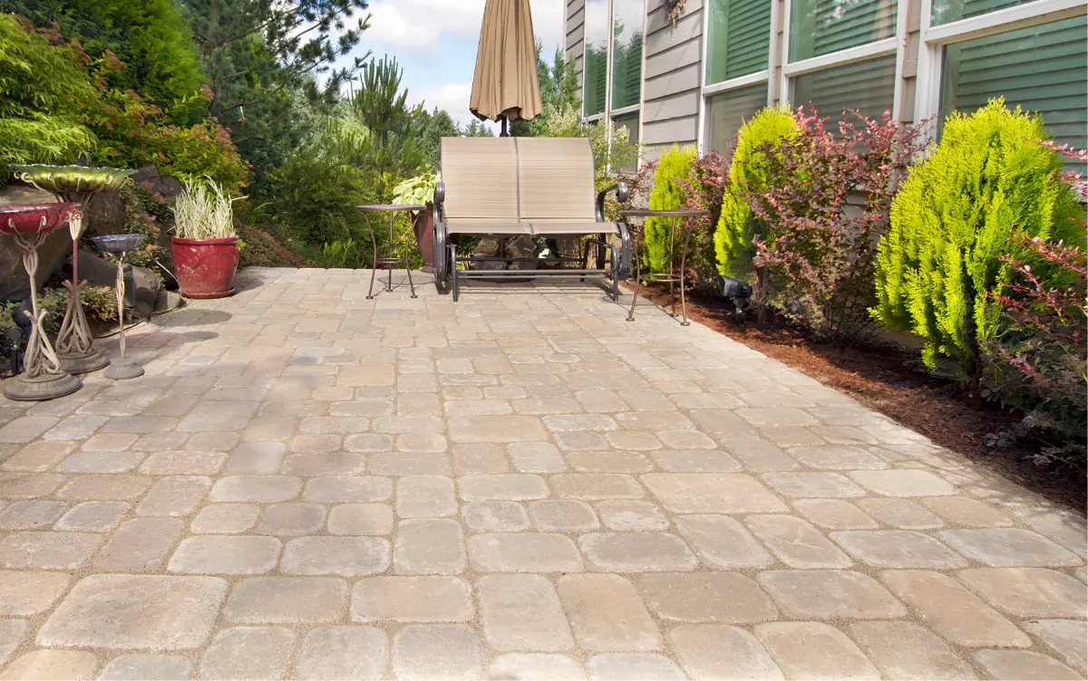 Outdoor patio with beige and gray interlocking pavers, a bench with a beige umbrella, two small side tables, and green shrubs along a house wall.