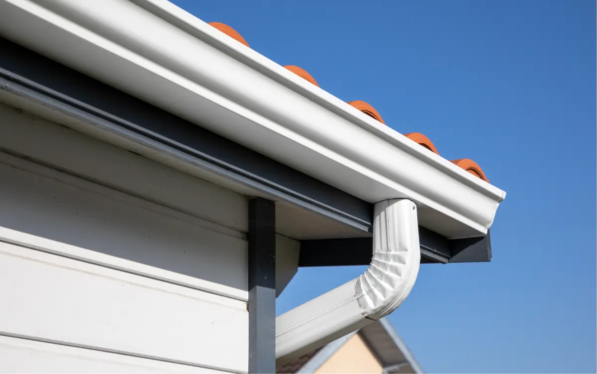 Close-up of a white rain gutter and downspout on the edge of a tiled roof against a clear blue sky.