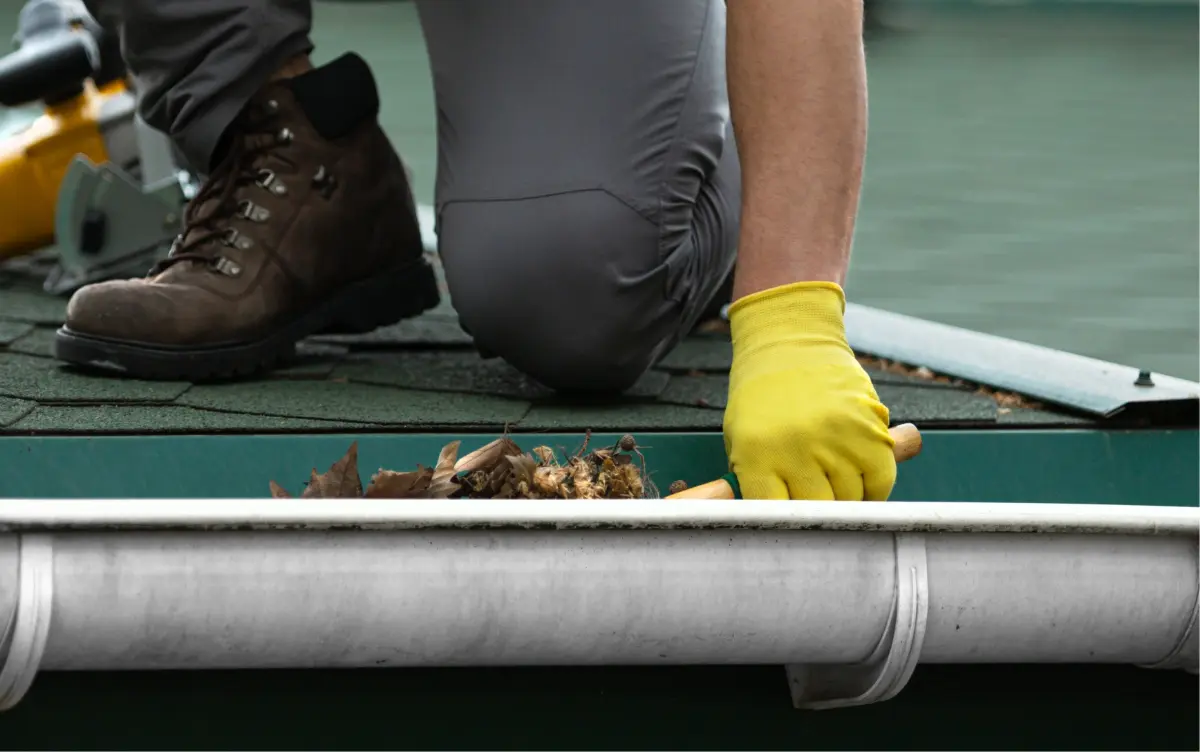 Person wearing yellow gloves and brown boots cleaning debris from a house gutter with a hand tool.