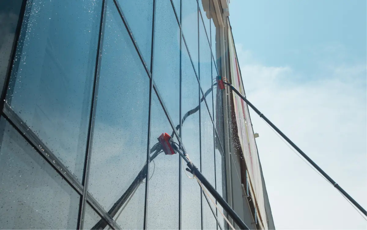 Two long-handled brushes cleaning a high glass building facade with water droplets on the windows.