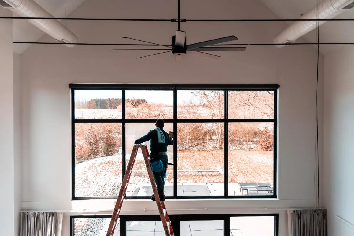 Person standing on a ladder cleaning a large indoor window with a scenic outdoor view.