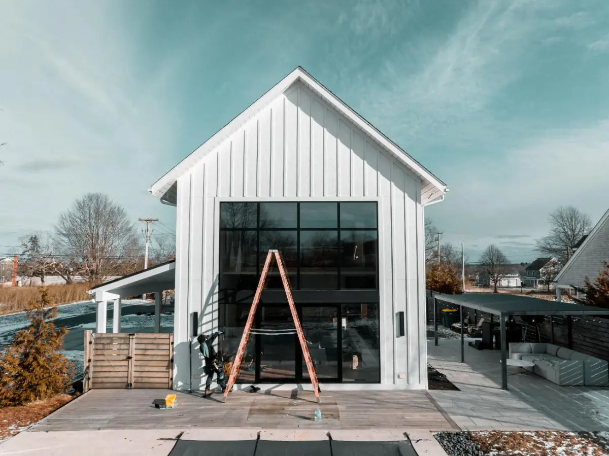 Modern white house with tall black-framed windows, a wooden deck, and a ladder leaning against the front.