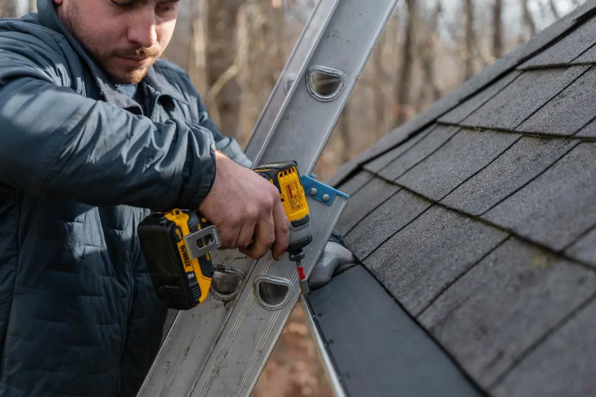 Man using a cordless drill to secure a metal gutter on the edge of a shingled roof while standing on a ladder.
