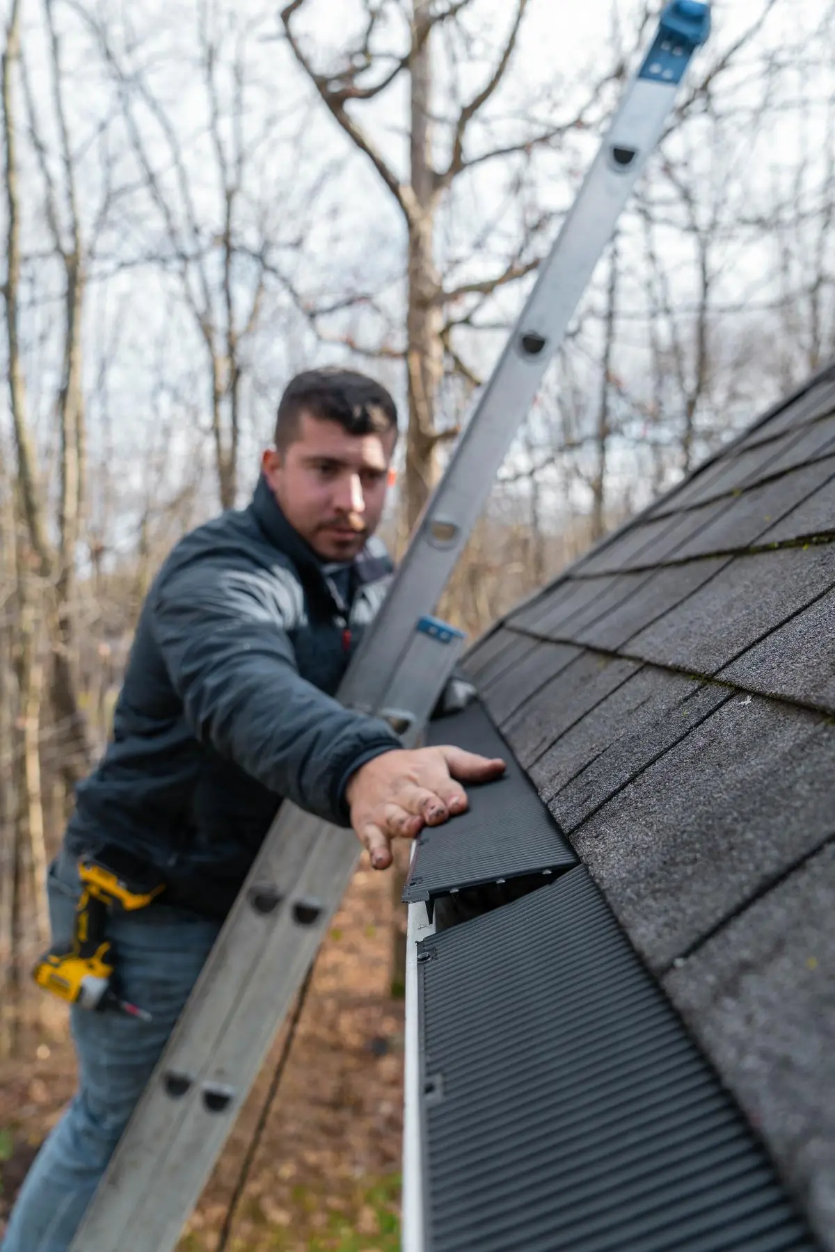 Man on a ladder installing a gutter guard on a roof in an outdoor wooded area.