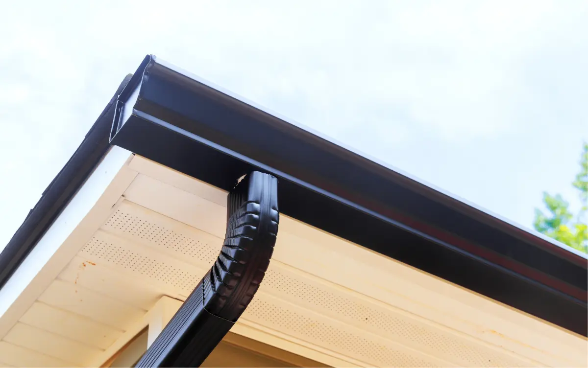 Close-up of a black gutter system installed on the corner of a beige house roof against a clear sky.