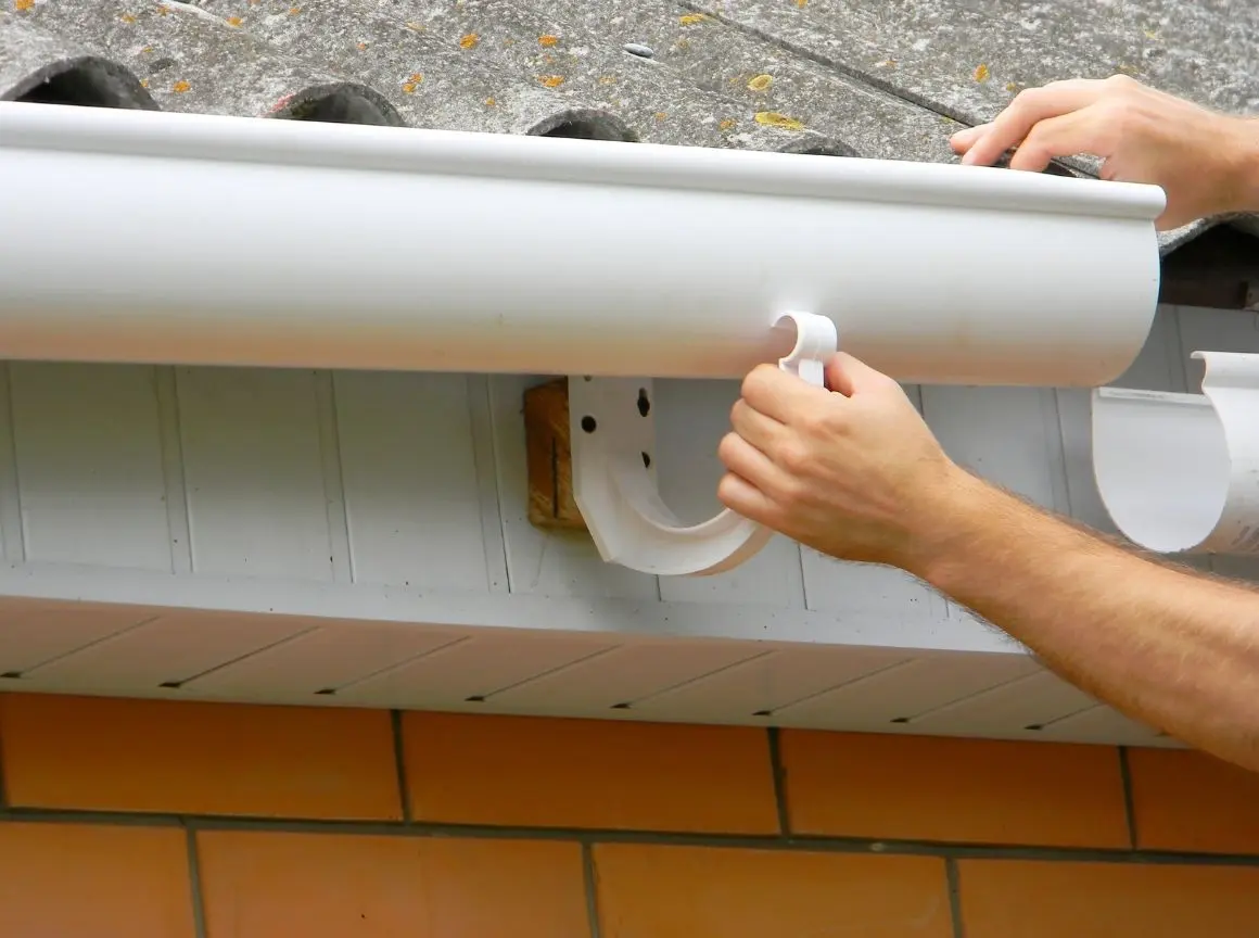 Person installing a white gutter onto a roof edge with visible roof tiles and brick wall below.