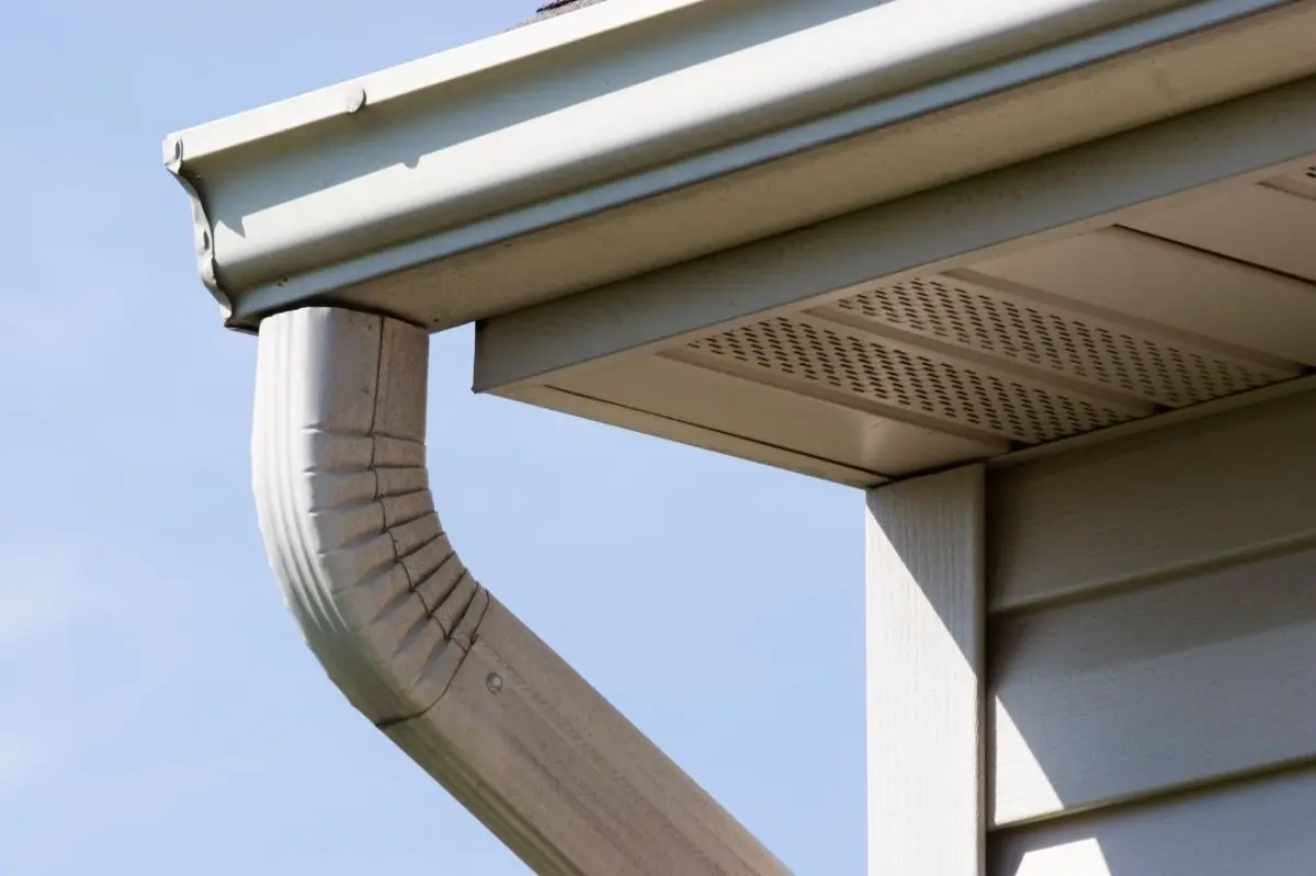 Close-up of a white house gutter and downspout connected to the roof against a blue sky.