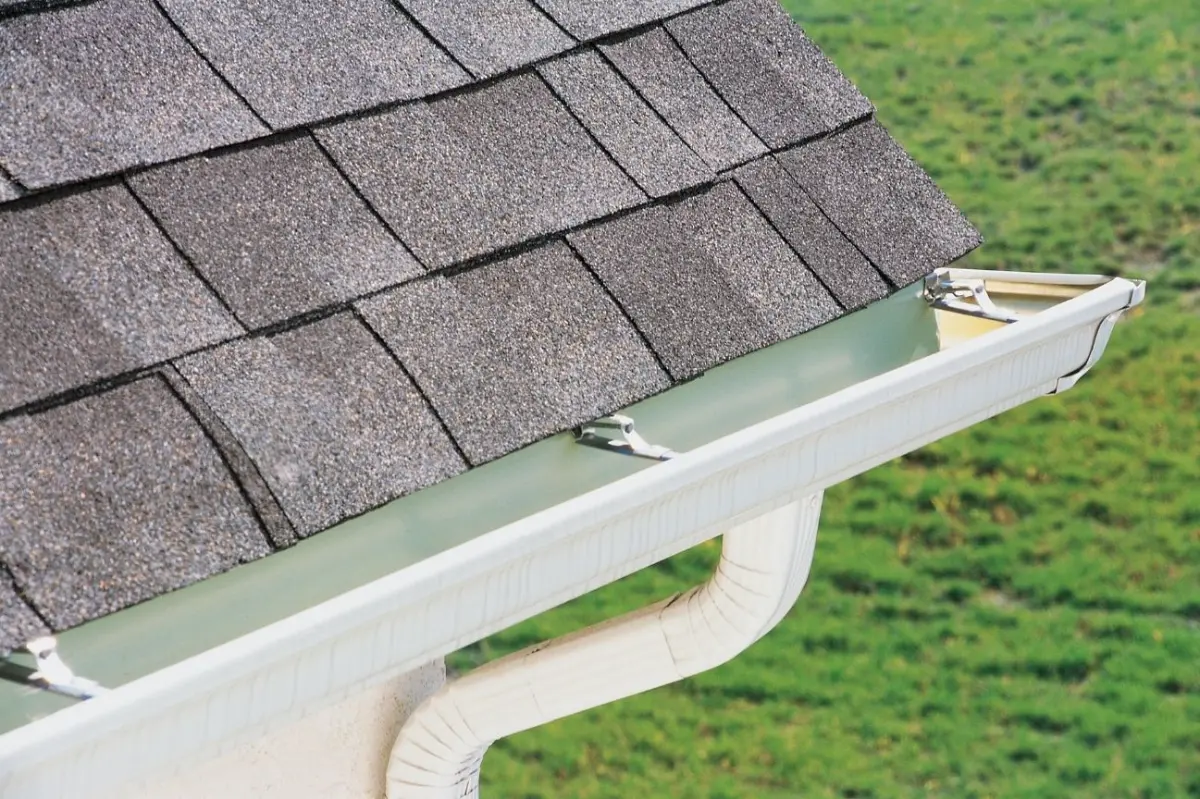 Close-up of a white rain gutter attached to a roof with gray asphalt shingles and green grass in the background.