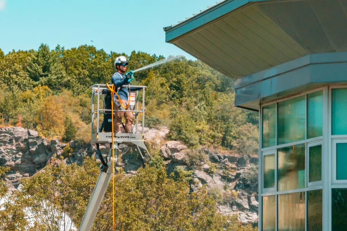 Worker wearing helmet and safety gear spraying water on a building’s exterior from an elevated lift platform.