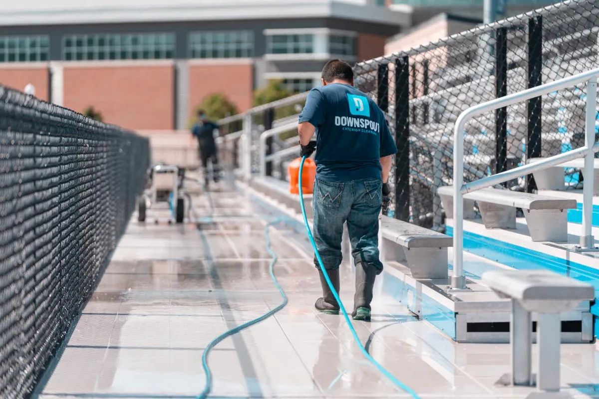 Person wearing rubber boots and a Downspout Exterior Cleaning shirt pressure washing bleachers walkway near chain-link fence.