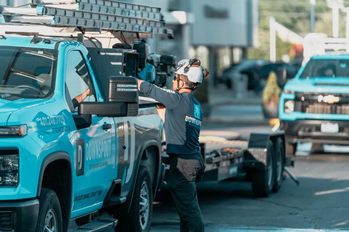 Worker wearing a helmet and safety glasses prepares equipment on a blue Downspout Exterior Cleaning truck with a trailer in an outdoor setting.