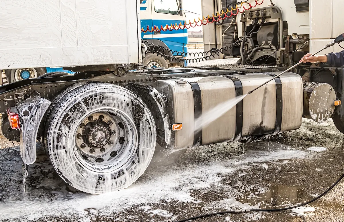 Person cleaning a large truck tire and fuel tank with a high-pressure water spray.