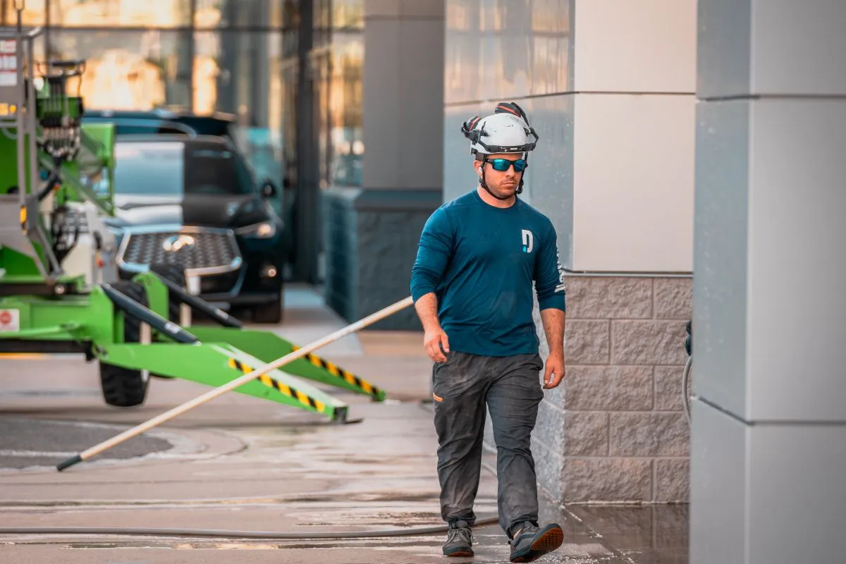 Construction worker wearing a white helmet and blue long-sleeve shirt walking outdoors near a building with a green machinery in the background.