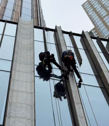 Two workers suspended by ropes cleaning the windows of a tall glass building.