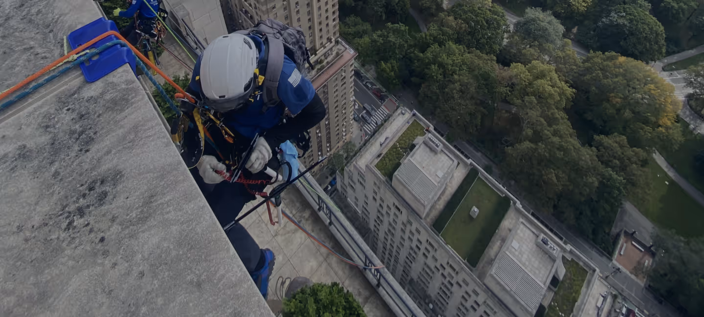 Person wearing safety gear and helmet rappelling down a tall building with trees and streets visible below.