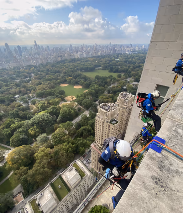 Workers wearing helmets and harnesses rappelling down the side of a tall building overlooking a city park with sports fields and dense urban skyline beyond.