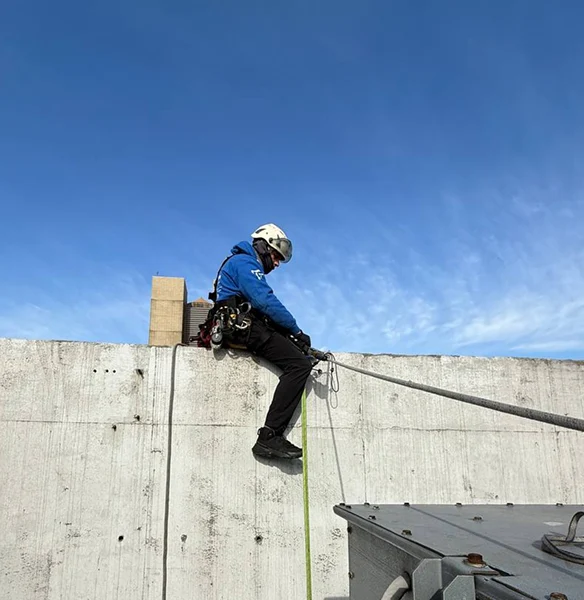 Worker wearing blue jacket and helmet rappelling down a concrete wall against a clear blue sky.