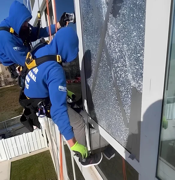Two workers in blue hoodies with safety harnesses repairing a shattered window on the side of a building while suspended by ropes.
