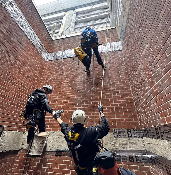 Three workers wearing helmets and safety harnesses conducting rope access work inside a brick-walled vertical shaft.